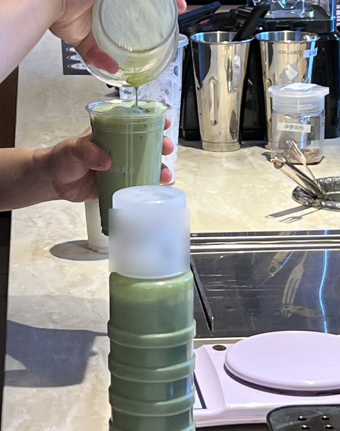 CHAGEEソウル店のカウンターで、秤にのせたカップに計量カップから抹茶の液を注いでいる手元。手前に朝に作った抹茶の液が入った白いボトル