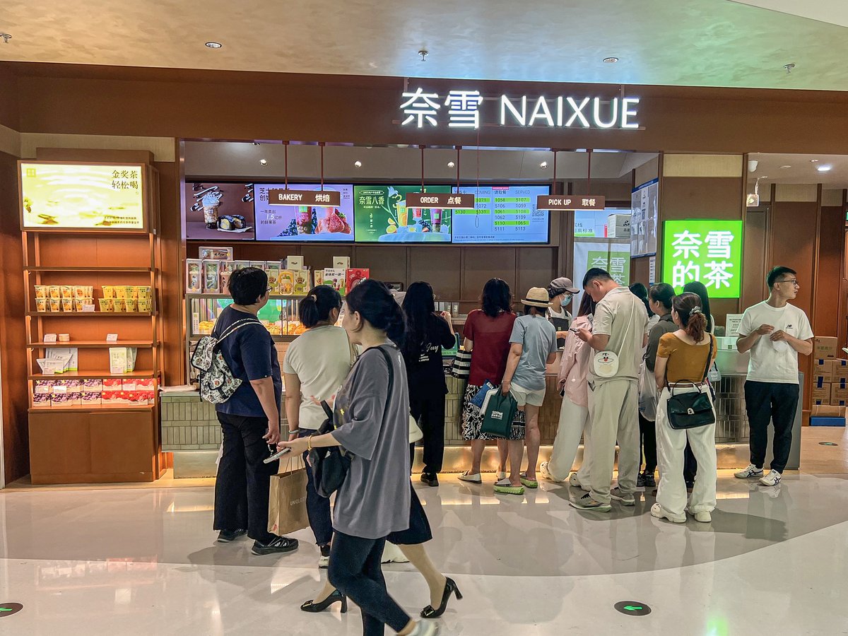 中国・北京の商業施設にある奈雪的茶（NAIXUE）の店舗前に並ぶ客の列。木目の壁に「奈雪 NAIXUE」の看板、緑の「奈雪的茶」のサイン、棚に並ぶ茶葉のギフト、注文と受け取りの画面