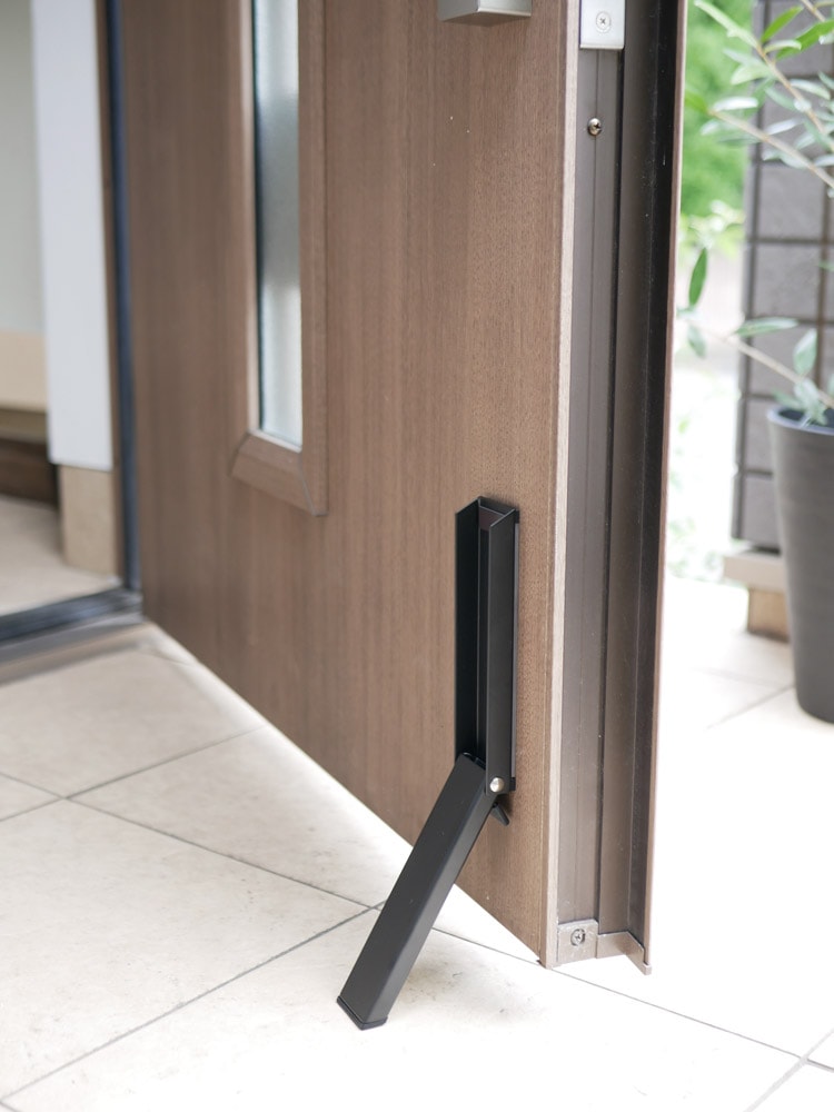tower（YAMAZAKI）のスマートマグネット折り畳みドアストッパーが、木製フローリングのスチールドア下部に設置されている様子。コンパクトな折り畳みデザインとマグネット部分が確認できる