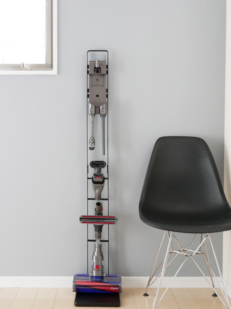 towerブランドのコードレスクリーナースタンドが木製床と棚、ランプと共にスタイリッシュに配置されている様子