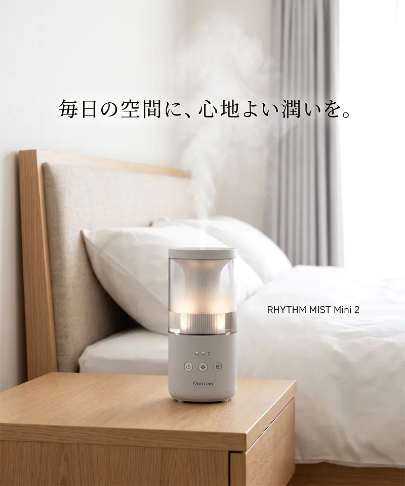 明るい寝室の木製ナイトスタンドに置かれた RHYTHM MIST Mini 2 プールレス加湿器。プールレス構造でミストと暖かな光を放出し、毎日の空間に心地よい潤いを与える。