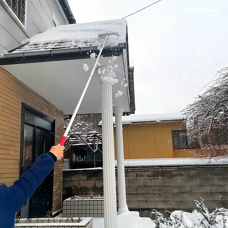 雪が積もった住宅の屋根から、長柄の雪下ろし道具を使って雪を除去している様子。屋根の端から雪を掻き出している。