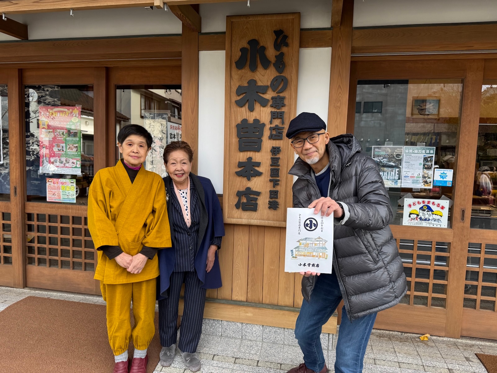 集合写真。小木曽商店下田本店前で看板を背景に撮影。左から小木曽商店女将、観音温泉の鈴木会長、ラジオパーソナリティの上柳昌彦さん。
