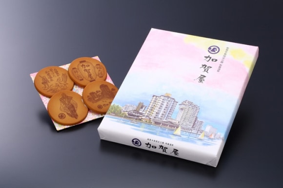 淡いピンクや水色の色使いで、ホテルの建物と水辺の風景が描かれた贈答用の箱と、和柄の敷物に並べられた4枚の加賀屋せんべい。