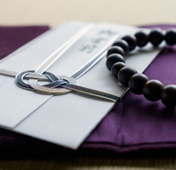 紫色の袱紗（ふくさ）の上に置かれた、黒白の水引が結ばれた不祝儀袋（のし袋）と数珠（念珠）。