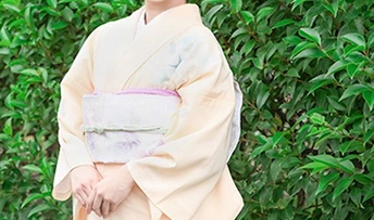 緑の木々を背景に、淡いクリーム色の夏着物（絽）を着た女性の腰から上のクローズアップ
